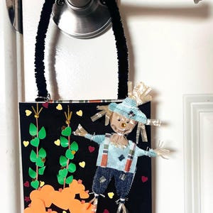 May include: A black rectangular wall hanging with a black fuzzy handle, featuring a fabric scarecrow with a blue hat and denim overalls, green paper vines, orange paper hearts, and a small label at the bottom reading MATHEW 9:35-38.