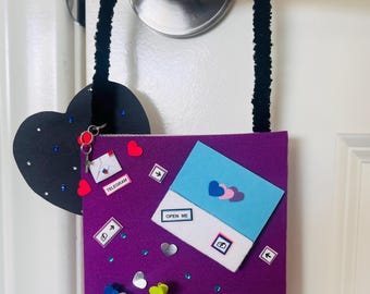 May include: A purple rectangular door hanger featuring a black fuzzy loop, a black heart cutout with blue rhinestones, and a silver cross charm. The surface displays a blue envelope labeled OPEN ME, a telegram icon, small heart cutouts, and a label reading PROVERBS 31: 25-26.