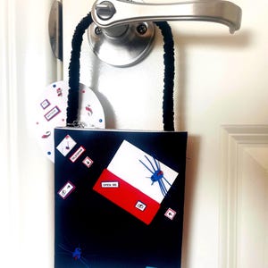 May include: A black rectangular hanging sign with a black fuzzy strap, featuring a white and red envelope graphic labeled OPEN ME, a blue spider, and a small blue tag at the bottom with the text PSALM 139: 1-14. A round white tag labeled ARTHUR hangs behind.