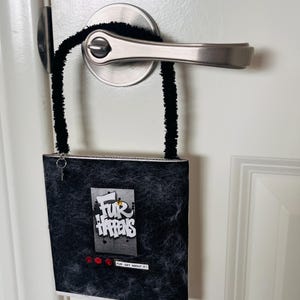 May include: A black door hanger with the text "Fur Happens" in white, grey, and yellow. The door hanger is attached to a door handle with black fuzzy pipe cleaners. The door hanger has three red buttons and a small key charm.