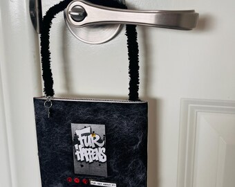 May include: A square black fuzzy sign hanging from a silver door handle by a black pipe cleaner loop. The sign features the white text "FUR HAPPENS" in a graffiti style, three small red circular accents, and a small label reading "FUR-GET ABOUT IT".