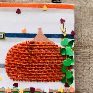 May include: A white fabric panel with orange and blue horizontal stripes featuring a textured orange burlap pumpkin applique. The design includes a green vine with heart-shaped leaves, small scattered heart cutouts, and a decorative border.