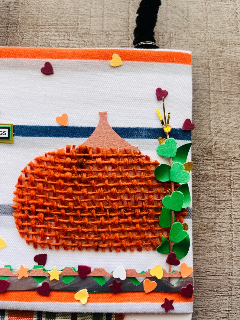 May include: A white fabric panel with orange and blue horizontal stripes featuring a textured orange burlap pumpkin applique. The design includes a green vine with heart-shaped leaves, small scattered heart cutouts, and a decorative border.