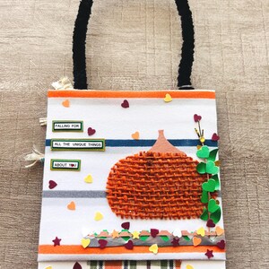May include: A rectangular hanging door note with a white background featuring orange and blue horizontal stripes. It displays a textured orange burlap pumpkin, green paper leaves, and small colorful hearts. Text reads: FALLING FOR ALL THE UNIQUE THINGS ABOUT YOU.