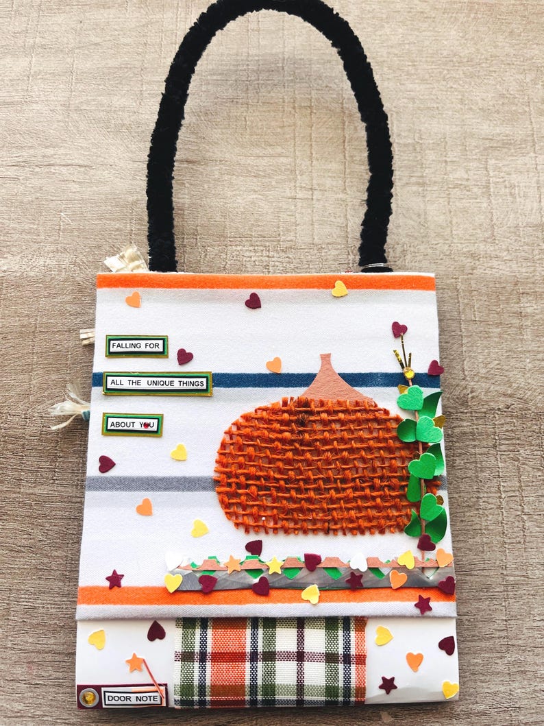 May include: A rectangular hanging door note with a white background featuring orange and blue horizontal stripes. It displays a textured orange burlap pumpkin, green paper leaves, and small colorful hearts. Text reads: FALLING FOR ALL THE UNIQUE THINGS ABOUT YOU.