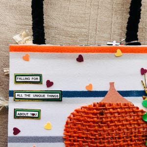 May include: A white fabric bag with black fuzzy handles featuring horizontal orange and blue stripes. The bag is decorated with a textured orange burlap pumpkin, small colorful heart cutouts, and three rectangular labels reading FALLING FOR, ALL THE UNIQUE THINGS, and ABOUT YOU.