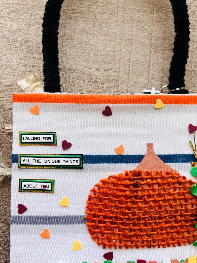 May include: A white fabric bag with black fuzzy handles featuring horizontal orange and blue stripes. The bag is decorated with a textured orange burlap pumpkin, small colorful heart cutouts, and three rectangular labels reading FALLING FOR, ALL THE UNIQUE THINGS, and ABOUT YOU.
