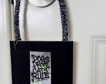 May include: A black rectangular hanging sign with a fuzzy black and white speckled handle. The sign features a graphic with the text "DOGS DROOL CATS RULE" in black graffiti-style lettering, three red circular accents, and a "CASE CLOSED" label.
