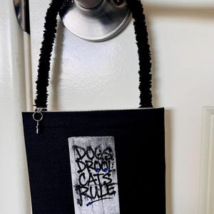 May include: A black fabric bag with a fuzzy black handle and a silver ring attachment. The front features a rectangular patch with the text DOGS DROOL CATS RULE, three small red circular accents, and a blue-bordered label reading I PREFER DROOL.