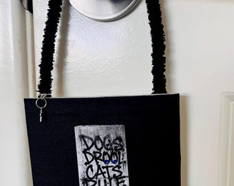 May include: A black fabric bag with a fuzzy black handle and a silver ring attachment. The front features a rectangular patch with the text DOGS DROOL CATS RULE, three small red circular accents, and a blue-bordered label reading I PREFER DROOL.