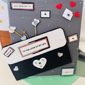 May include: A handmade grey card holder decorated with paper hearts, arrows, and the text "LOVE NOTE FOR YOU" and "TO THE LOVE OF MY LIFE". The card holder has a black and white envelope design with the words "OPEN ME".