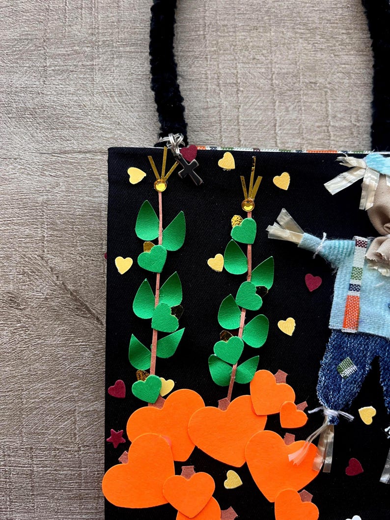 May include: A black fabric tote bag featuring a decorative applique design. The scene includes two green vine stalks with heart-shaped leaves, orange heart-shaped pumpkins, and a small scarecrow figure wearing a blue shirt and denim pants.