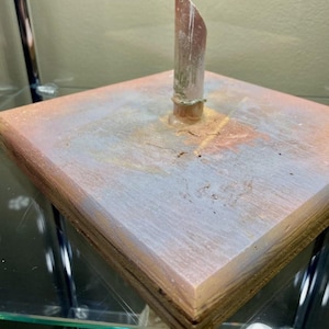 May include: A square, multi-layered wooden base with a copper-colored finish and a vertical, cylindrical, copper-colored object rising from the center. The base has a distressed, aged appearance with a mix of pink, gray, and brown tones.
