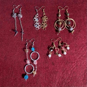 May include: Five pairs of handmade earrings on a red background. The earrings feature various designs, including beaded, chain, and hoop styles. Colors include silver, gold, blue, pink, and clear beads. Some earrings have dangling charms.