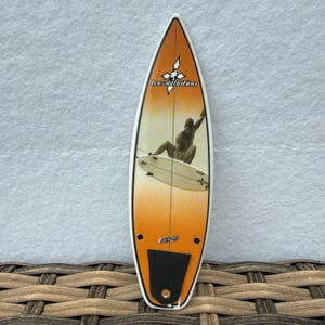 May include: A surfboard with an orange and yellow gradient finish featuring a graphic of a person surfing. The board displays the text Local Motion at the top and includes a black traction pad near the bottom with a single screw hole visible.