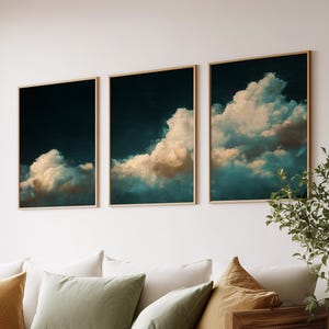 Puede incluir: Tres impresiones artísticas enmarcadas que representan un dramático paisaje de nubes con un cielo azul oscuro y nubes blancas y verde azulado esponjosas. La obra de arte se exhibe sobre un sofá blanco con cojines decorativos en tonos verdes y dorados.