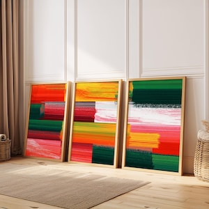 May include: Three framed abstract art prints. Each print features a colourful, textured design with horizontal brushstrokes in shades of red, orange, yellow, green, and pink. The frames are a light wood colour.