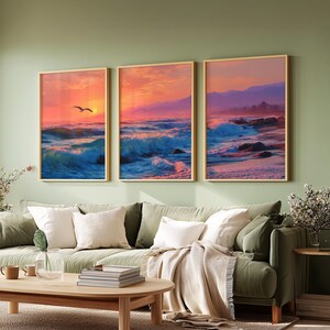 May include: Three framed wall art pieces depicting a vibrant ocean sunset. The artwork features shades of blue, orange, and pink, with crashing waves and a seagull. The frames are a light wood color, adding a natural touch to the coastal theme.