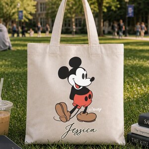 May include: A beige canvas tote bag featuring a classic Mickey Mouse design with the name "Jessica" in script. The bag has a black and red Mickey Mouse graphic and is ideal for carrying books and other items.