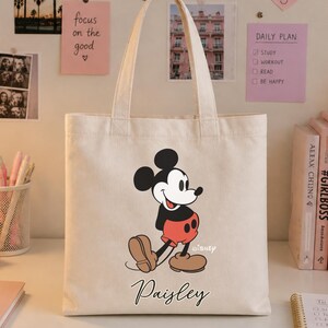 May include: A cream-colored tote bag featuring a classic Mickey Mouse graphic in black, red, and tan. The bag has a personalized name, "Paisley," in script font. The bag is on a white desk with stationery and books.