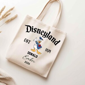 May include: Cream-colored tote bag featuring the Disneyland logo with Donald Duck. The bag includes the text "EST 1934" and "DONALD Ember 2026". The bag has two cream-colored handles.