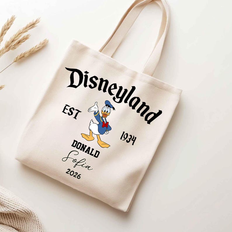 May include: Cream-colored tote bag featuring the Disneyland logo with Donald Duck. The bag includes the text "EST 1934" and "DONALD Sofia 2026". The bag has two handles and is made of a canvas-like material.
