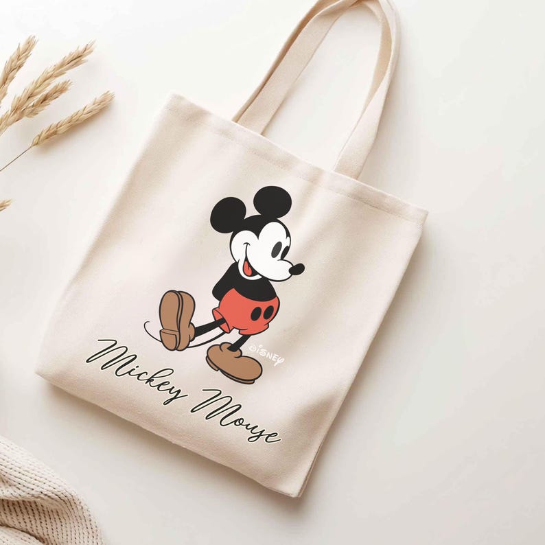 May include: Cream-colored tote bag featuring a classic Mickey Mouse graphic. The design includes Mickey in his signature red shorts, black top, and brown shoes. The words "Mickey Mouse" are written in a cursive font below the character.