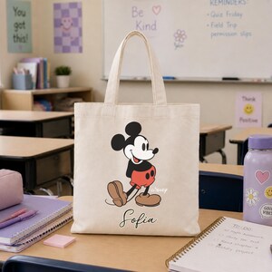May include: A beige canvas tote bag featuring a cartoon image of Mickey Mouse in his classic outfit. The bag has a black handle and the name "Sofia" printed in cursive below the character. The background shows a classroom setting.