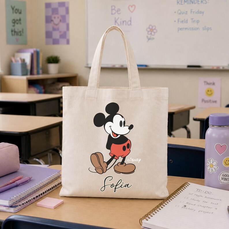 May include: A beige canvas tote bag featuring a cartoon image of Mickey Mouse in his classic outfit. The bag has a black handle and the name "Sofia" printed in cursive below the character. The background shows a classroom setting.