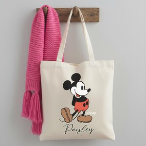 May include: Off-white canvas tote bag featuring a Mickey Mouse graphic in black, red, and brown. The bag has long white handles and the name "Paisley" printed in black script. A pink knitted scarf hangs on a wooden hook.