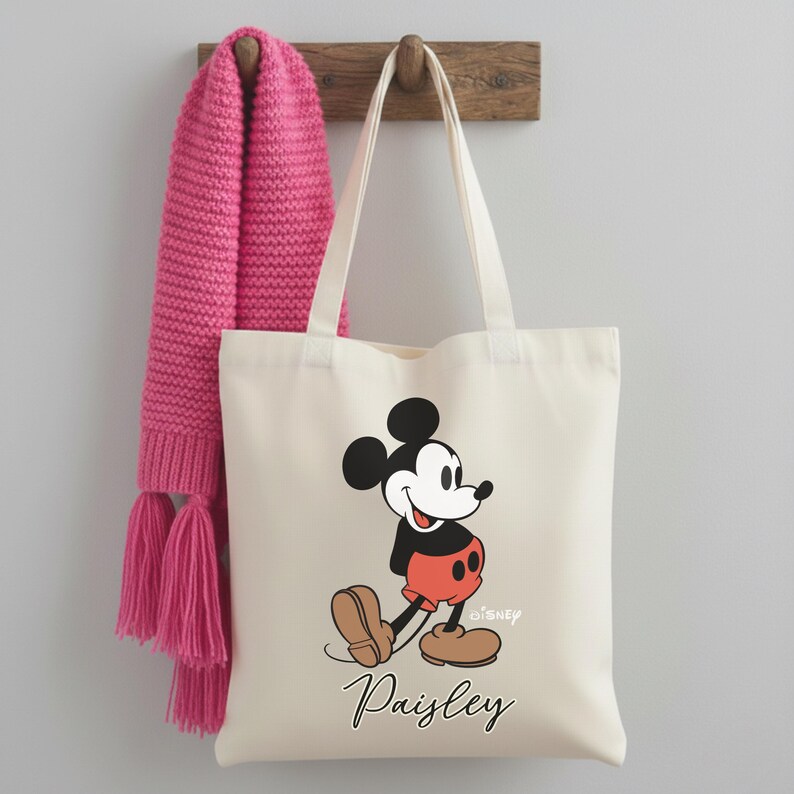 May include: Off-white canvas tote bag featuring a Mickey Mouse graphic in black, red, and brown. The bag has long white handles and the name "Paisley" printed in black script. A pink knitted scarf hangs on a wooden hook.