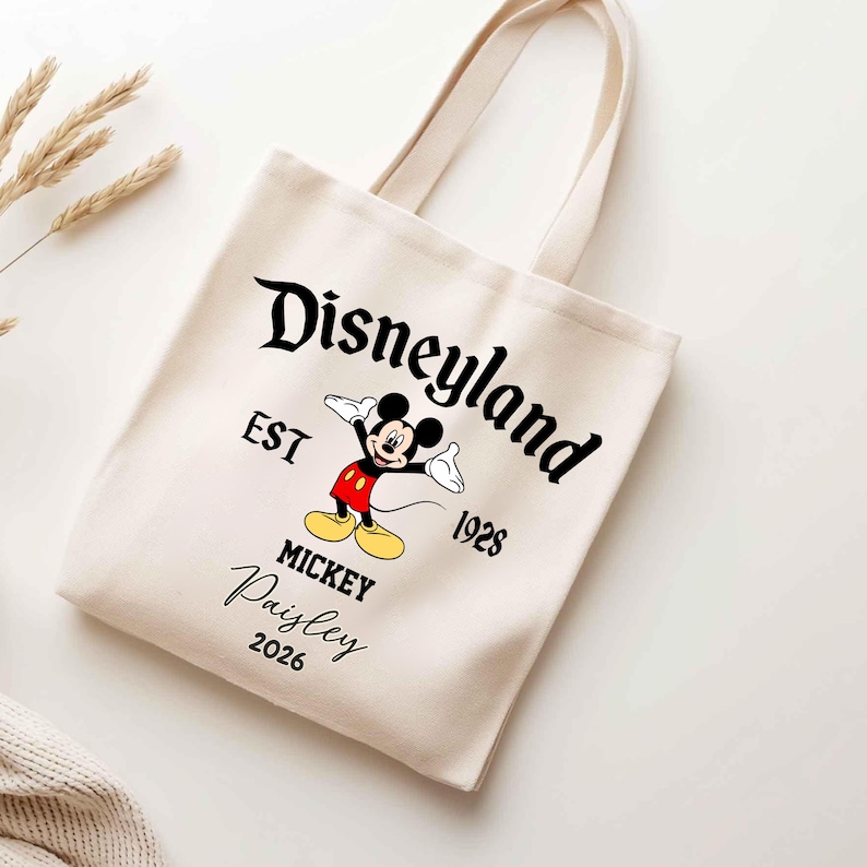 May include: Cream-colored canvas tote bag featuring the Disneyland logo with Mickey Mouse. The design includes the text "EST 1928" and "Mickey Paisley 2026". The bag has two handles.