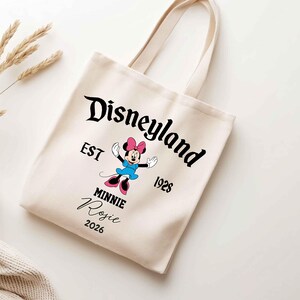 May include: Cream-colored tote bag featuring the Disneyland logo and a Minnie Mouse graphic. The bag includes the text "EST 1928" and the name "Rosie" with the year "2026". The bag has two handles.