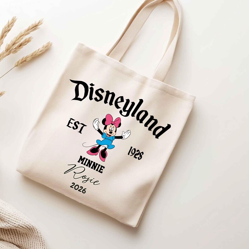 May include: Cream-colored tote bag featuring the Disneyland logo and a Minnie Mouse graphic. The bag includes the text "EST 1928" and the name "Rosie" with the year "2026". The bag has two handles.