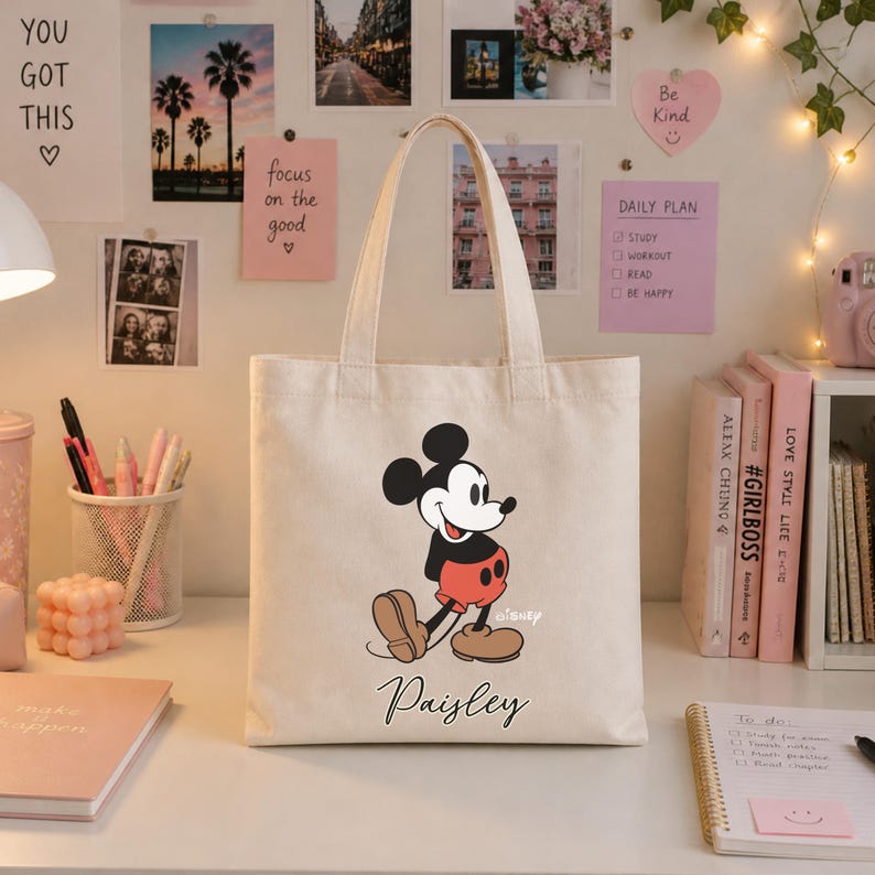 May include: A cream-colored tote bag featuring a classic Mickey Mouse graphic in black, red, and tan. The bag has a personalized name, "Paisley," in script font. The bag is on a white desk with stationery and books.
