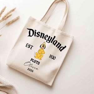 May include: Cream-colored tote bag featuring the Disneyland logo in black script, with "EST 1930" and a yellow Pluto graphic. The name "Olivia" and the year "2026" are also printed on the bag. The bag has two cream-colored handles.