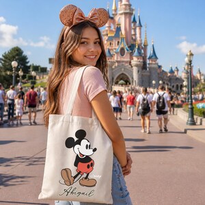 May include: A beige canvas tote bag featuring a classic Mickey Mouse illustration in black, red, and white. The bag has the name "Abigail" in script and the Disney logo. The background shows a theme park with a castle.
