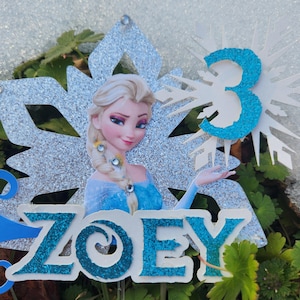 May include: A glittery cake topper featuring a cartoon image of Elsa from Frozen, with the name "ZOEY" and the number "3". The design includes a silver snowflake and is set against a backdrop of green foliage.