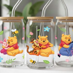 Könnte beinhalten: Drei klare Glastrinkbecher mit Bambusdeckeln und Glasstrohhalmen. Jeder Becher zeigt eine Grafik von Winnie Puuh, der eine andere Figur umarmt: Ferkel, Tigger oder I-Aah. Die Designs enthalten kleine bunte Blumen, Blätter und Bienen.