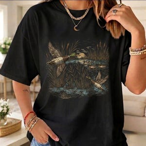 Puede incluir: Camiseta negra con una ilustración detallada de patos volando sobre un estanque, rodeados de juncos. El diseño utiliza tonos de marrón, verde y azul para representar una escena natural, mostrando aves acuáticas.