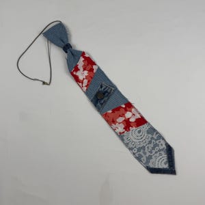May include: A handmade denim tie with patchwork design. The tie features sections of blue denim, red floral fabric, and white lace. A small denim pocket with a button is sewn onto the tie. A thin, dark-colored cord is attached to the tie.