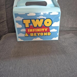 May include: A light blue cardboard treat box with a handle, featuring a white cloud pattern on a blue sky background. The front displays the yellow text TWO, a red banner with the word INFINITY, and the yellow text & BEYOND in a bold font.