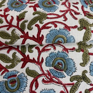 May include: A white fabric featuring a repeating floral pattern with light blue petals, dark red stems, and olive green leaves. The textile has a visible woven texture and is folded to show the continuous print across the surface of the material.