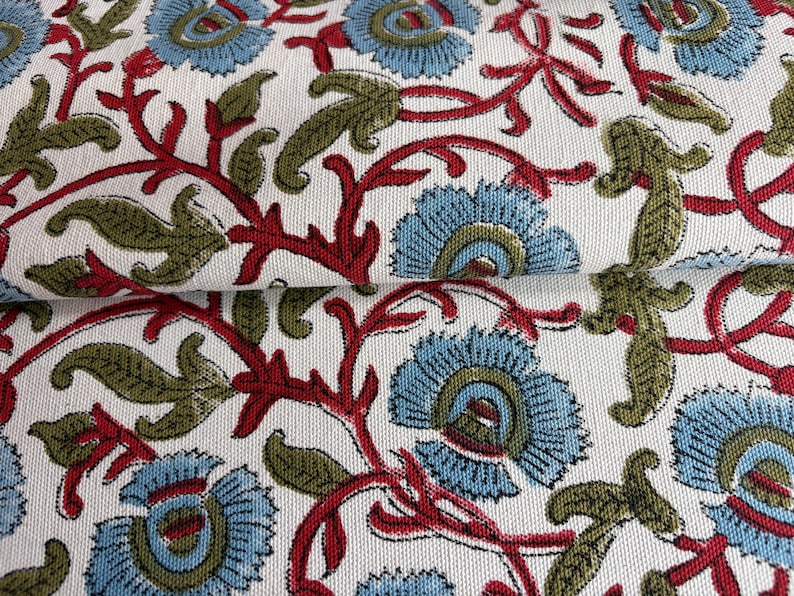 May include: A white fabric featuring a repeating floral pattern with light blue petals, dark red stems, and olive green leaves. The textile has a visible woven texture and is folded to show the continuous print across the surface of the material.