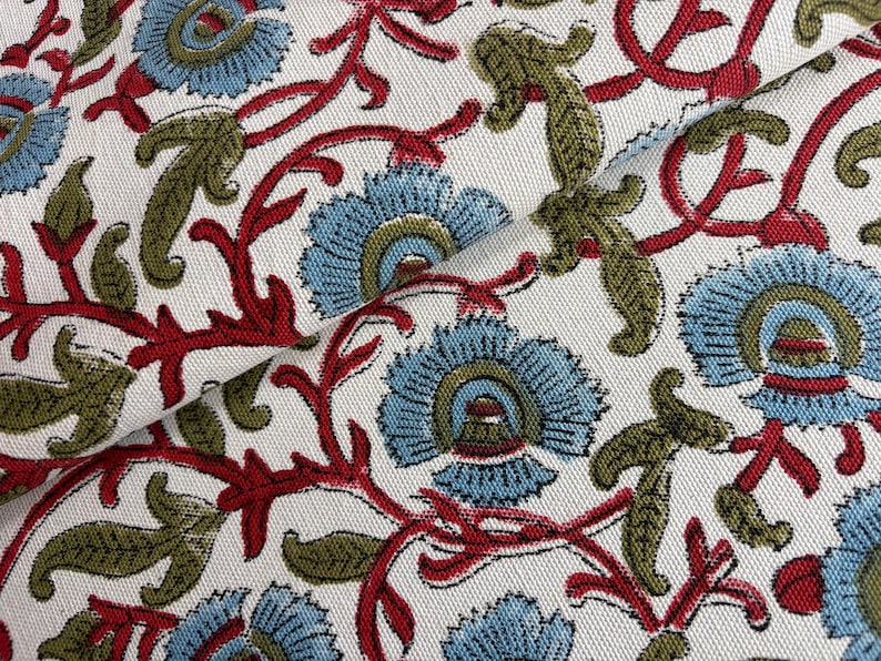 May include: A white fabric featuring a repeating floral pattern with light blue petals, olive green leaves, and dark red stems. The textile has a visible woven texture and a dense arrangement of botanical motifs across the entire surface area.