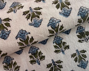 May include: Close-up of a white fabric with a repeating floral pattern. The design features blue flowers and green leaves. The fabric is folded, showing the texture and pattern variation.