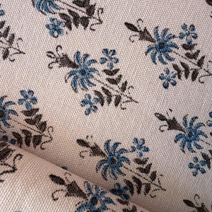 Puede incluir: Primer plano de una tela color crema con un patrón repetido de diseños florales azules y marrones. Las flores tienen una apariencia detallada y estilizada. La tela está ligeramente doblada, mostrando su textura.