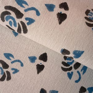May include: Close-up of a cream-colored fabric with a repeating floral pattern. The flowers are stylized, with black petals and blue accents. The fabric is folded, showing the texture and design.