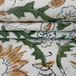 May include: Close-up of folded fabric with a repeating floral pattern. The design features green, tan, and black botanical elements on a cream-coloured background. The fabric has a textured appearance.