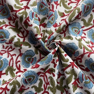 May include: A white fabric featuring a repeating floral pattern with light blue petals, olive green leaves, and red stems. The textile is gathered in the center, creating folds that highlight the intricate botanical print across the surface.